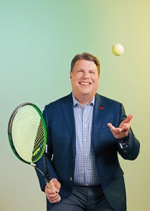 Dean Jamie Prenkert holding tennis racket with his left hand throwing up a tennis ball.