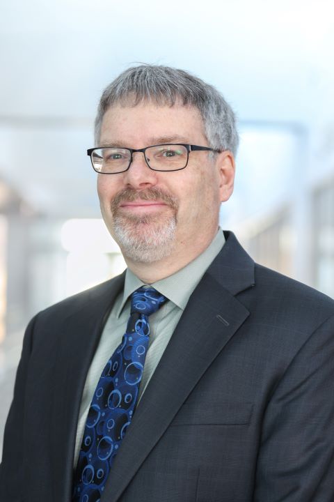 Headshot of Professor John Budd wearing a dark suit in front of blurred lighted hallway
