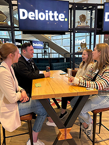 Four young adults sitting at a wooden table in what appears to be a Deloitte office or event space. 