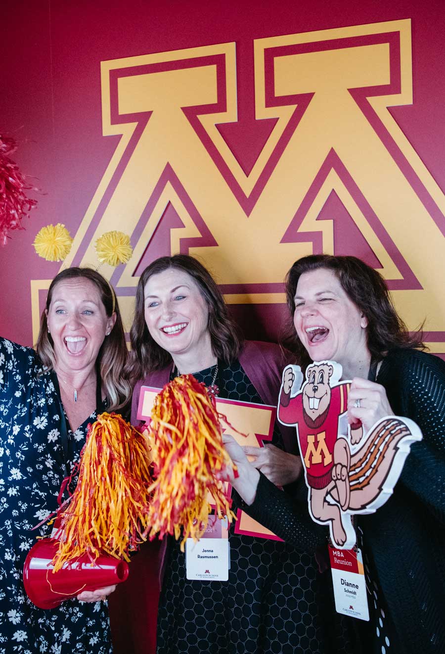 Three people pose for a photo in front of the "M" logo. They are holding props such as pompoms and a Goldy Gopher to show their school spirit.