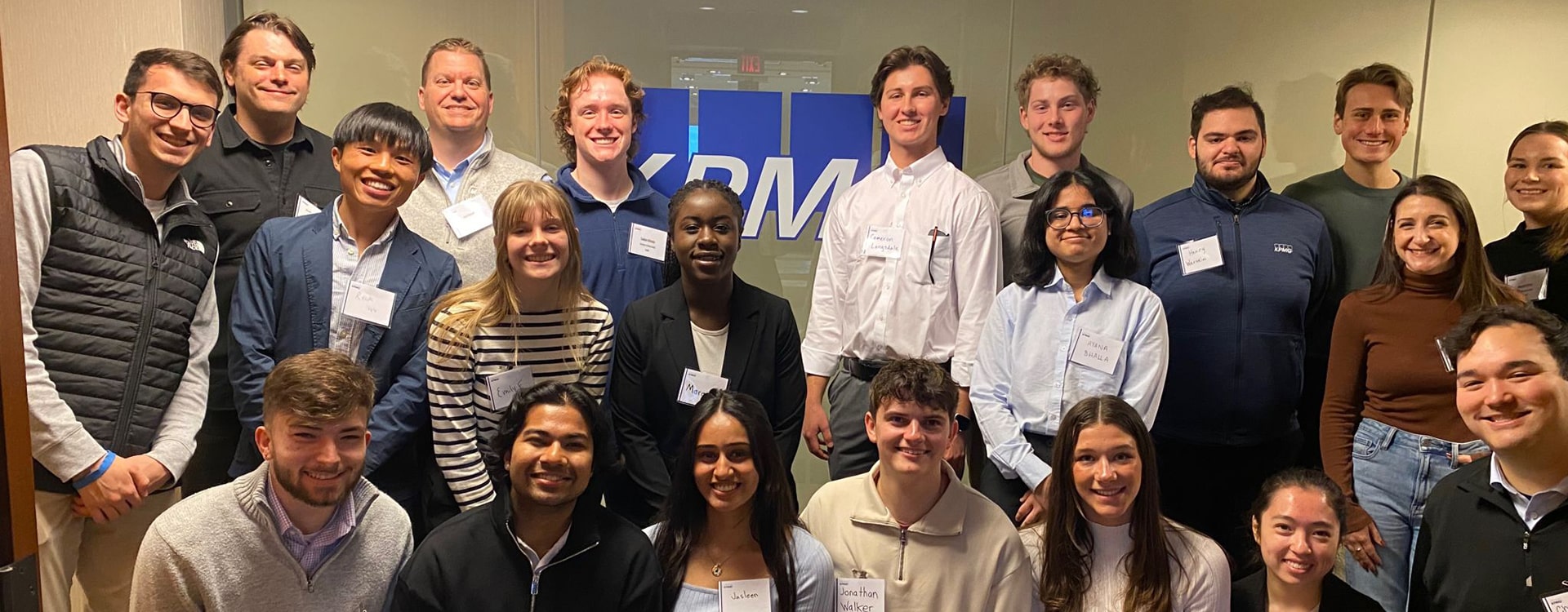 A group photo of approximately 20-25 students, standing together in front of a wall with a "KPMG" logo visible. 