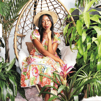 Shayla Owodunni sits in a wooden chair surrounded by large green plants.