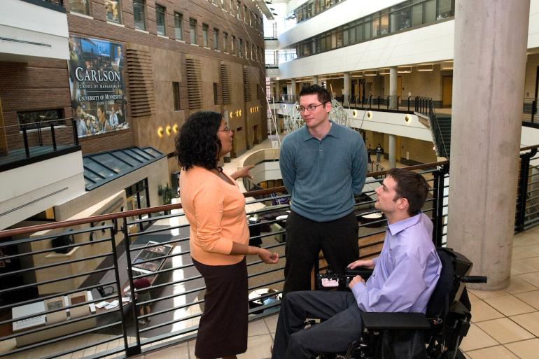 A woman and a man are standing with a man using a wheelchair, having a conversation in the Carlson School of Management