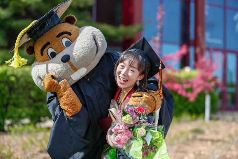 Goldie the Gopher, wearing a graduation cap and gown, has their arm around a smiling woman wearing a graduation cap and golding flowers