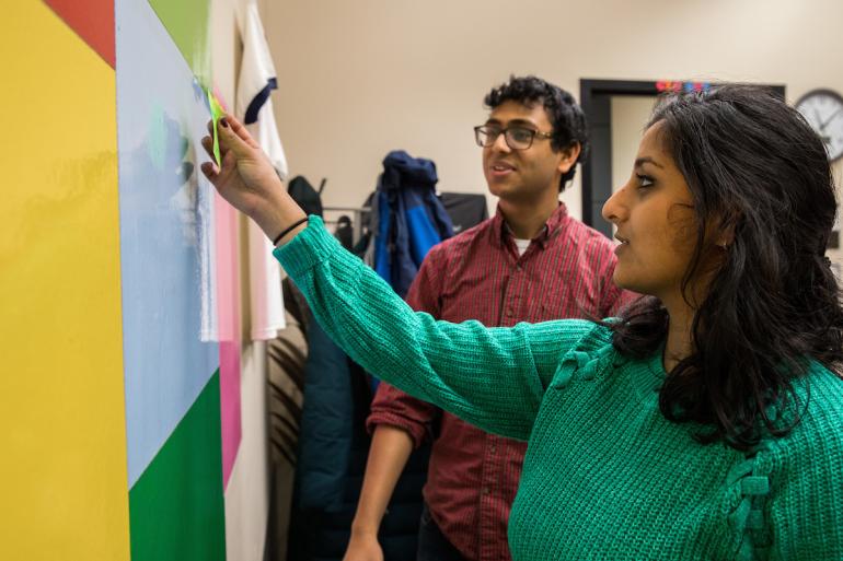 Two Ventures Enterprise students put up post-it notes on colorful paper taped to the wall