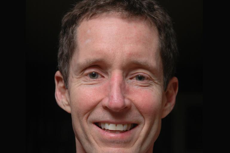 A Pgoto of Jeff Bailey from the shoulders up, smiling at the camera against a stark black background. He is wearing a pink collared shirt.