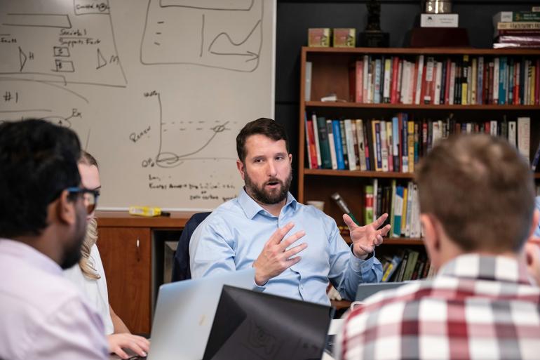  A bearded man in a light blue shirt gestures while speaking to a group in an office setting. 