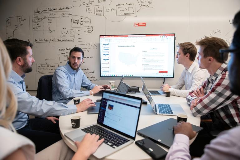 A diverse team of professionals in a meeting room. They are gathered around a table with laptops, discussing a presentation displayed on a large screen. 