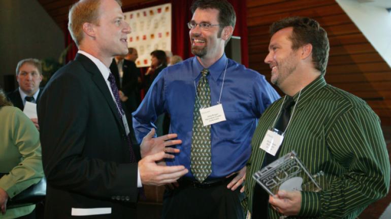 Two men smiling holding an award speak to another man.