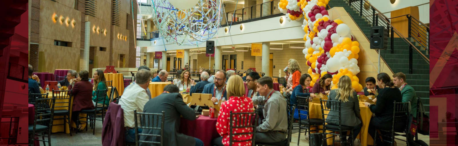 Alumni sitting at tables in the Carlson School atrium, with balloons and other maroon and gold decorations.