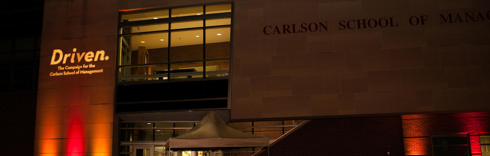 Exterior image of the Carlson School of Management at night with maroon and gold up lights.