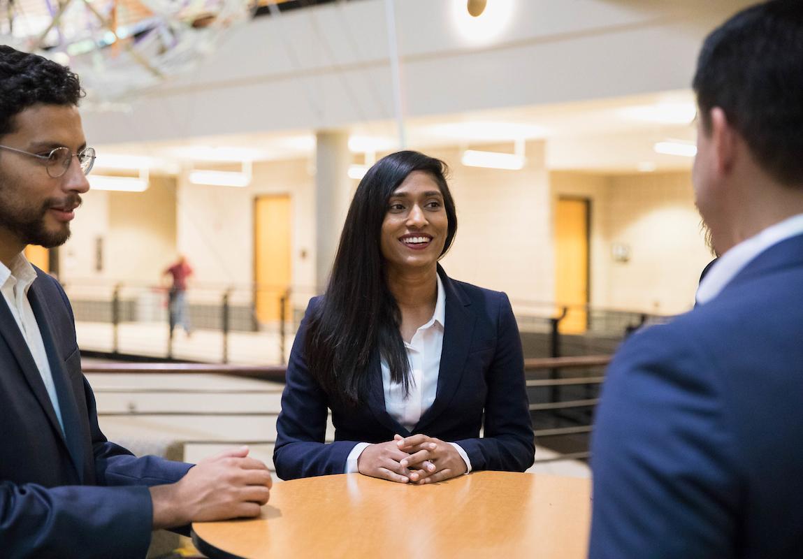 The focus is on a smiling woman with long dark hair in the center, wearing a navy blazer and white shirt. She is facing two men, one to her left wearing glasses and another partially visible to her right. They are standing around a round table in a large open space with visible hallways and doors in the background.