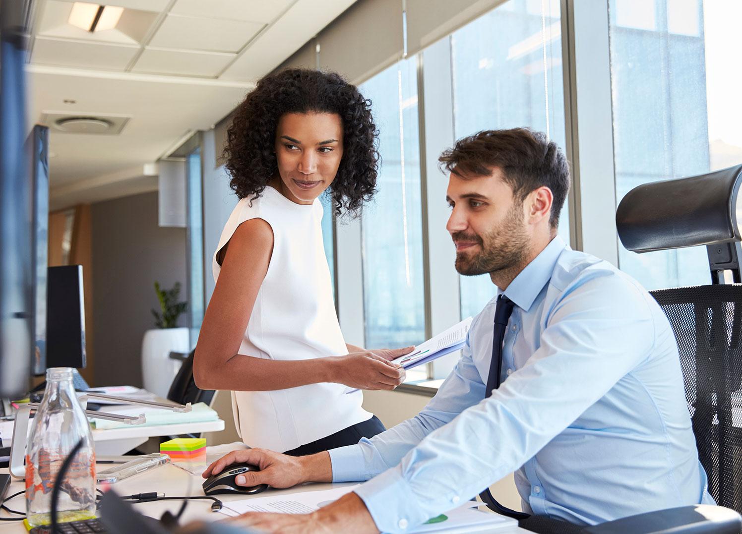 woman sitting on edge of desk while man reviews charts on computer monitor