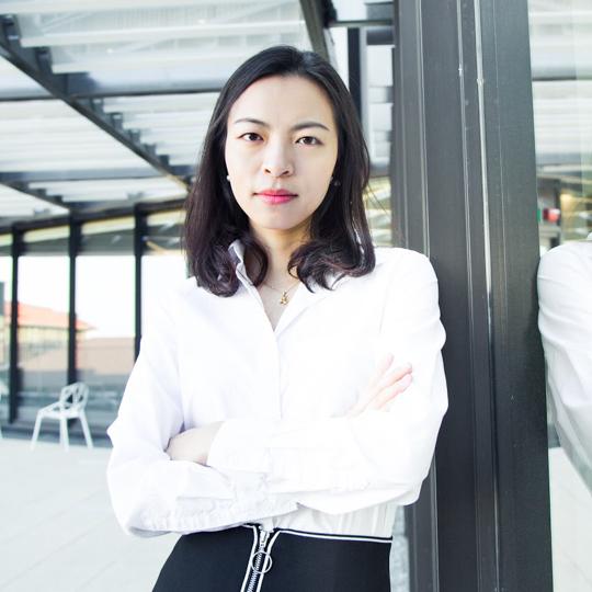 Mindy Zhang Xiaolan is pictured from the waist up, her arms crossed and her face is neutral. She is wearing a white, collared, button down shirt and a black skirt with white detailing.