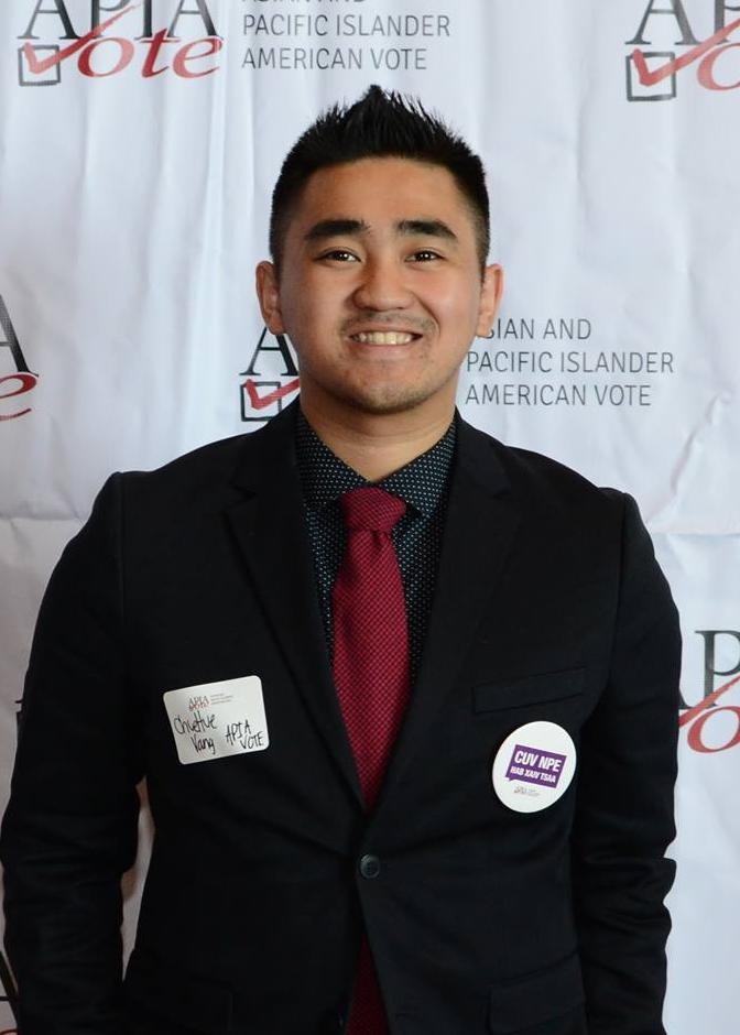 Chuehue is wearing a black suit with a red tie and standing in front of a white and red American Pacific Islander Vote backdrop in this photograph.