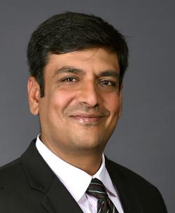 A picture of Vikrant Vig from the chest up, smiling closed-lipped at the camera. He is wearing a black suit jacket with a white shirt and red, black and gray striped tie.