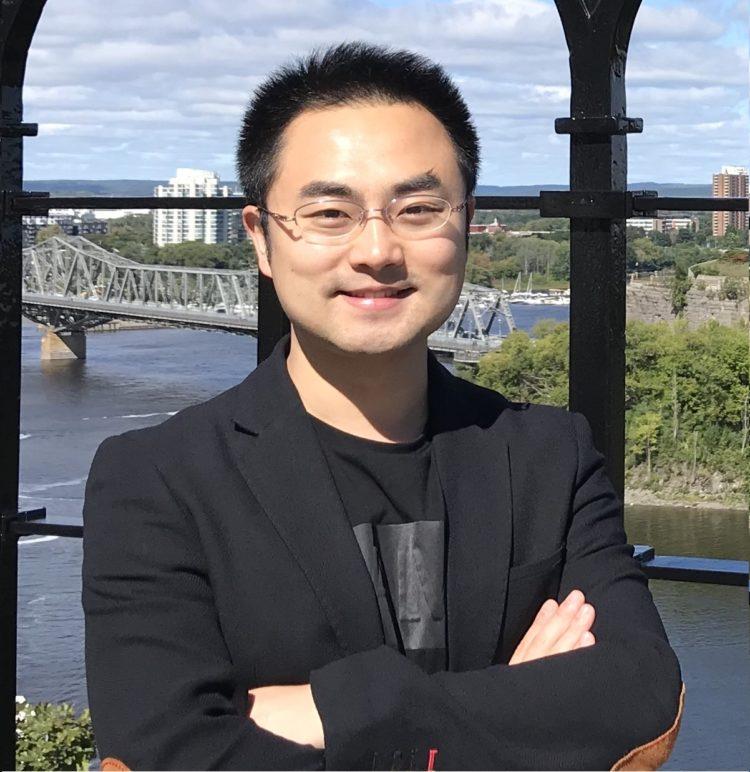 Ye Li is pictured from the chest up. He is wearing a black blazer over a black graphic t-shirt. He is smiling and wears glasses.