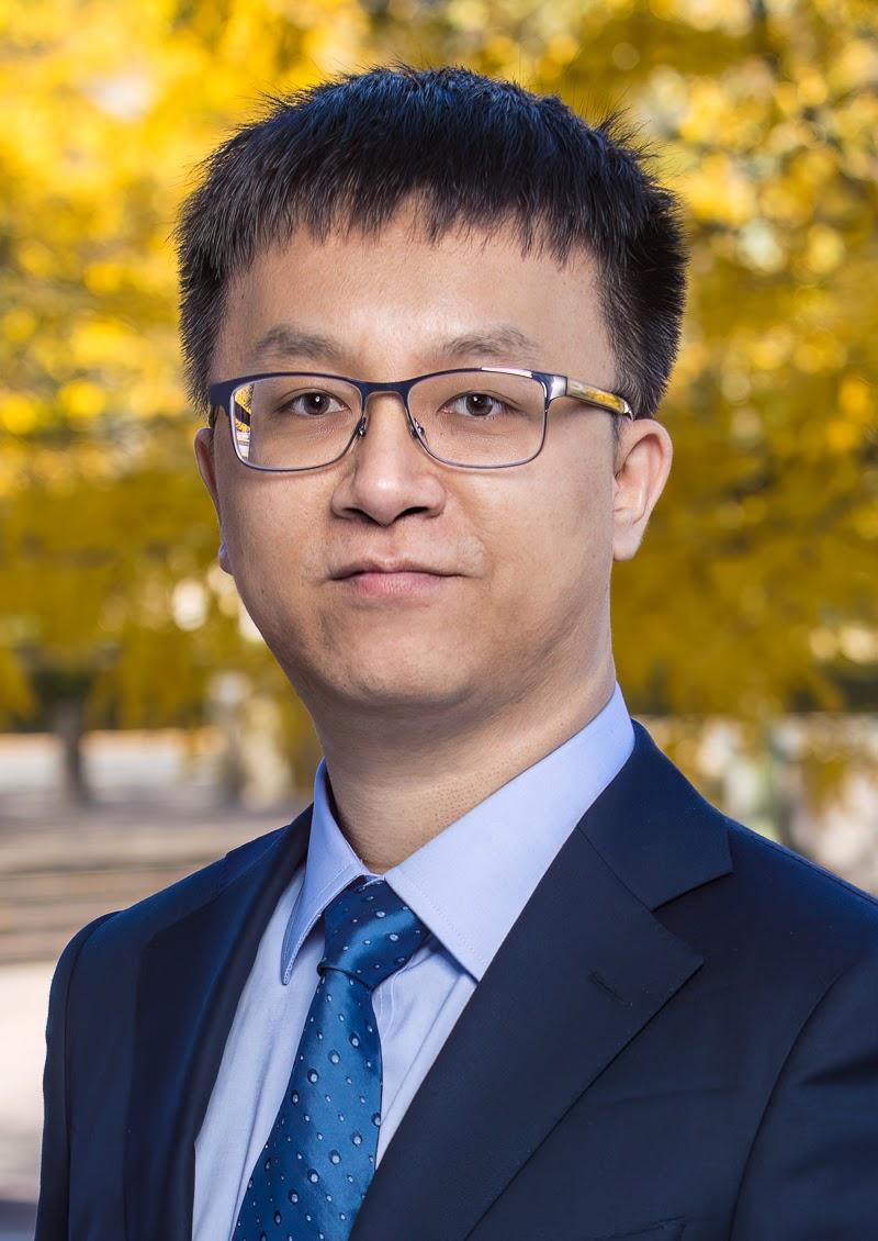 Yucheng Yang is pictured from the chest up. He is wearing a dark blue suit coat over a light blue shirt and royal blue tie. He has a neutral expression and wears glasses.