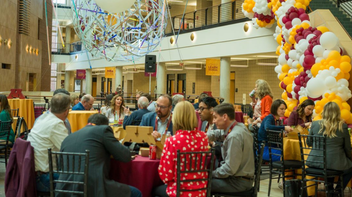 Alumni sitting at tables in the Carlson School atrium, with balloons and other maroon and gold decorations.