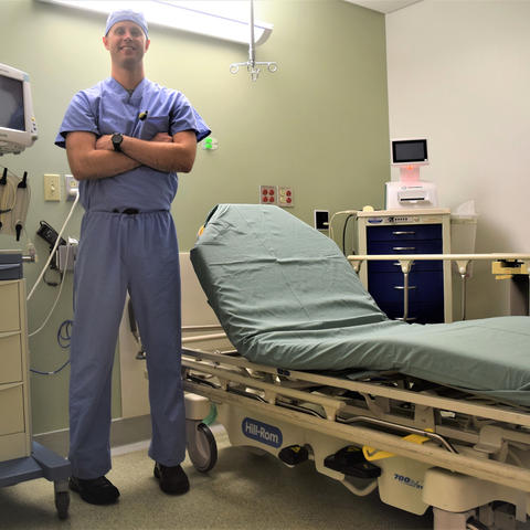 Thomas Kaster standing next to empty medical examination bed in scrubs