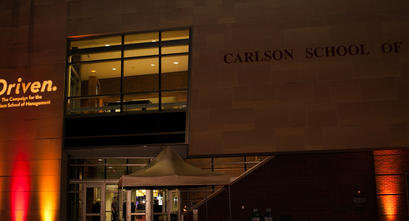 Exterior image of the Carlson School of Management at night with maroon and gold up lights.
