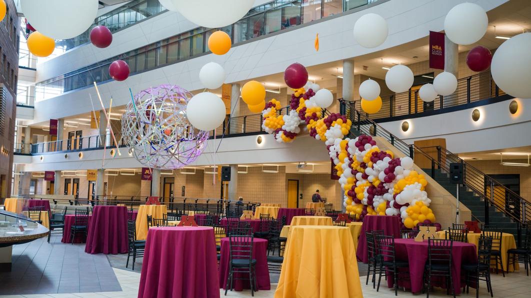 A decorated Carlson School atrium. Tables have maroon and gold tablecloths, and there are maroon, gold, and white balloon decorations.