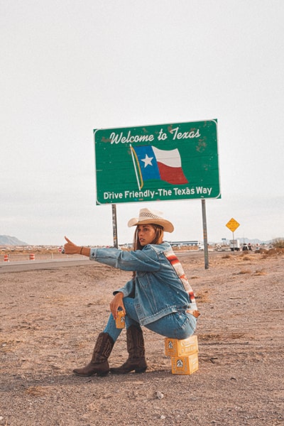 Young woman wearing a jean jacket, books and a cowboy hat, sitting on a box in front of a "Welcome to Texas" sign.